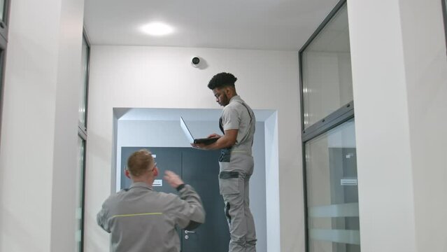 Two multi ethnic installers uniform sets up security cameras in office corridor using laptop. Upset African American worker looks at camera. Concept of surveillance systems and privacy. Quick zoom in