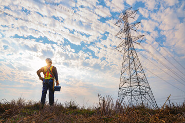 Technician stands carrying a toolbox to a high-voltage pylon wearing an operator fall arrester with hooks for safety harnesses. Concept of maintenance work on height equipment at high voltage pylons.