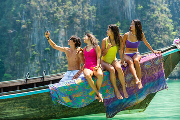 Group of Asian man and woman friends using mobile phone taking selfie together during travel on boat passing island beach lagoon in sunny day. People enjoy and fun outdoor lifestyle on summer vacation