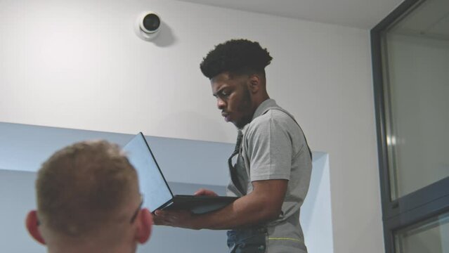 Two multi ethnic installers in uniform sets up security cameras in office hallway using laptop. They talk, check CCTV cameras in computer program. Concept of surveillance systems and privacy. Zoom in.