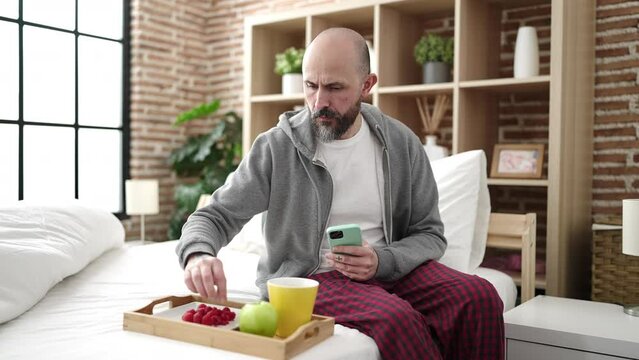 Young bald man make photo to breakfast sitting on bed at bedroom