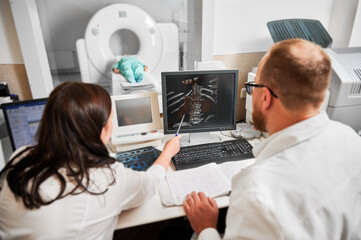 Medical computed tomography or MRI scanner. Back view of two doctors sitting, working at computer,...