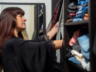 Woman standing in front of hanger rack and trying to choose an outfit in the morning. Redhead girl wearing a nightgown chooses clothes in her closet for working. Home wardrobe with folded shirts.