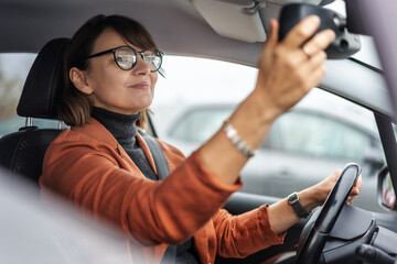 Adult caucasian businesswoman in glasses adjusting the rearview mirror while sitting at the wheel of a car