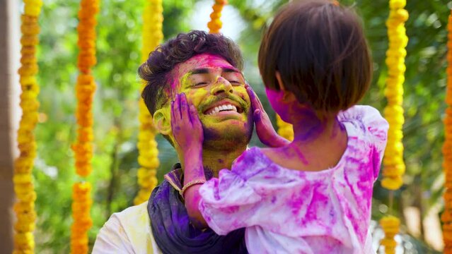 kid applying holi colors to caring fathers face on flower decorated background - concept of holi celebration, togetherness and relationship bonding.