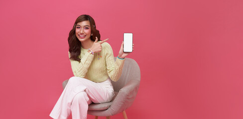 Happy young Asian woman showing empty screen mobile phone. While her sitting on chair isolated on...
