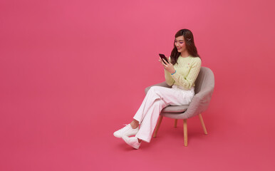 Happy young Asian woman using mobile phone. While her sitting on chair isolated on pink studio copy space background.