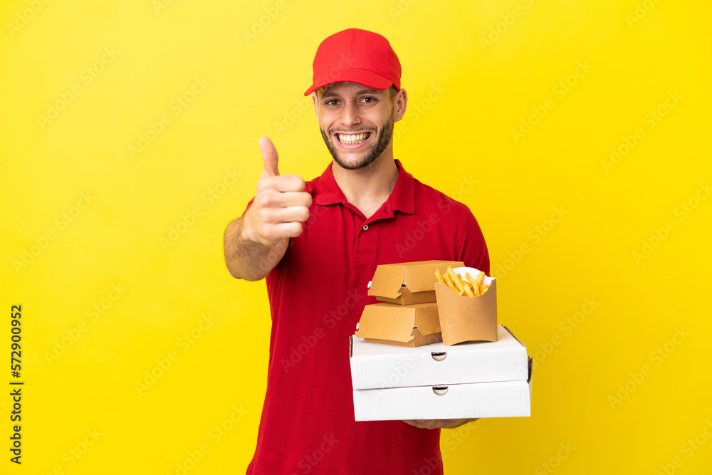 Wall mural pizza delivery man picking up pizza boxes and burgers over isolated background with thumbs up because something good has happened