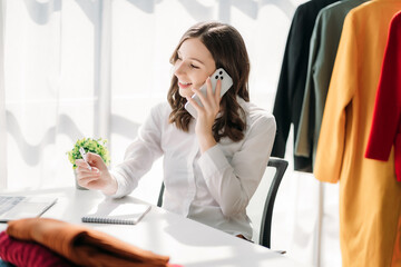 Fashion designer woman talking smart phone and using laptop with digital tablet computer in modern studio in morning light.