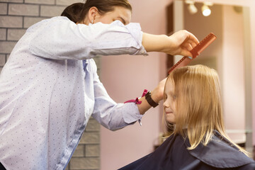 Barber woman make fashionable hairstyle for little blond girl child in barbershop. Hairdresser makes hairdo for baby in barber shop. Hairstyle, hair salon, beauty concept. Copy advertising text space