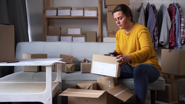 Small online business owner preparing small cardboard parcel for postage deliveries in home office working on smartphone in warehouse. Small business ecommerce