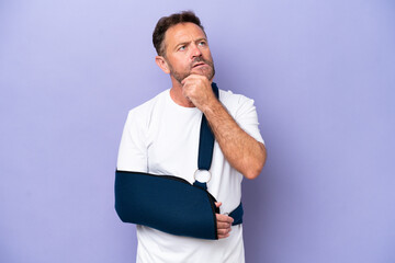 Middle age caucasian man with broken arm and wearing a sling isolated on purple background having...