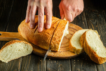 Close-up of a chef's hands with a knife cutting wheat bread on a kitchen cutting board. Bread on...