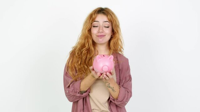 Young redhead woman unhappy and holding a piggybank over isolated background