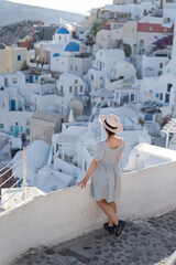 Fototapeta na wymiar Tourist woman in blue dress with hat relaxing at sea view in the traditional village Oia. Walking Santorini tour, Greece, during her summer crouise travel. European tourism attraction in Greece.