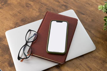 Clean Desk Essentials: Wooden desk with a plant, white paper, glasses, book, pen,office supplies, and phone with blank white screen.