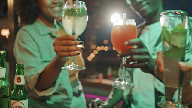 Selective focus medium close-up of three friends sitting at bar counter clinking glasses with cocktail drinks