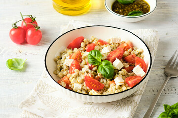 Warm salad of boiled pearl barley, ripe tomatoes, feta cheese with pesto sauce in a white plate on a light wooden background.