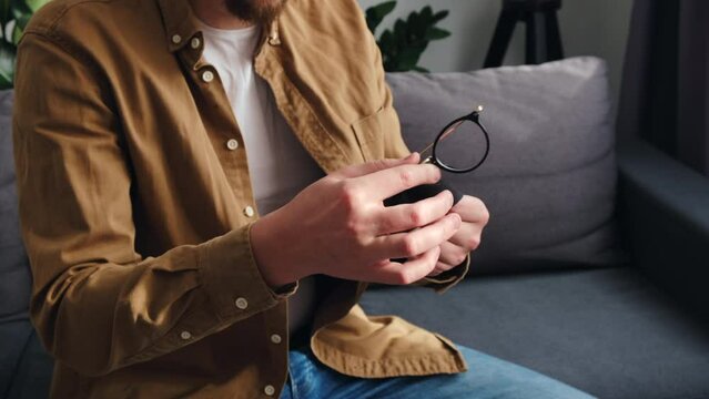 Close up of young caucasian male cleaning eyeglasses with small black cloth sitting on comfortable sofa in living room at home. Cleaning glasses concept. Selective focus