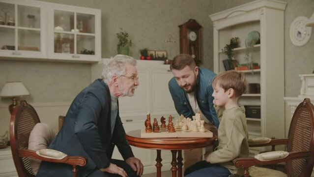 Three generations of men from the same family play chess at home. The grandson makes a move by rearranging a piece on the chessboard. Dad and Grandpa are watching. A family hobby. 