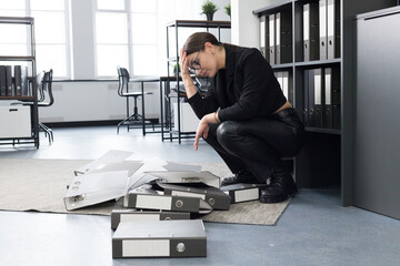 Young woman in the office drops a stack of folders and crouches next to them, holding her head and contemplating a tough work day.