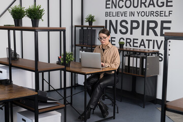 A young female office worker with glasses and a beige shirt sitting at her computer, pondering her work. Her desk features a wooden table, a notebook, a small plant, and various stationery items.