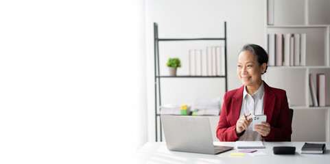 Senior Asian businesswoman in bright red suit using her smartphone while working in the modern office room.