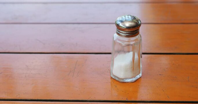 Pepper and salt shaker on wooden table, woman takes pepper to add to food. Close-up, only fingers and two pots are visible, slow motion hand-held shot.