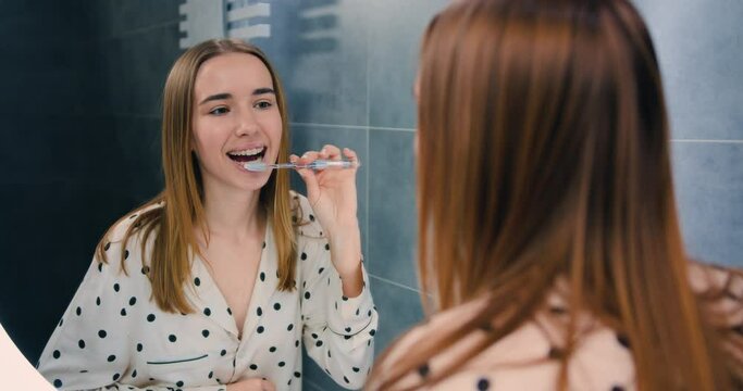 Young smilling woman with braces brushing teeth in modern bathroom and looking at reflection in mirror. Oral hygiene. Teen girl cleaning.
