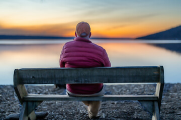 Winter scene of a man with red jacket in the middle of a bench looking out over a lake at sunset. ...