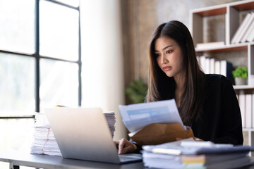 Concentrated young beautiful businesswoman working on laptop and in have documents on hand at bright modern office