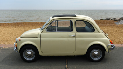 Classic Cream Coloured Fiat 500 parked on seafront promenade beach and sea in background. - obrazy, fototapety, plakaty