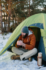 A young bearded man rests in the winter mountains near a tent. A man-traveler with a beard in a cap and a warm jacket warms up by drinking hot tea or coffee after a hike. Travel, lifestyle