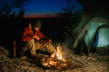 A bearded young man rests by the night campfire in the winter forest. The concept of survival and hiking in the wild.