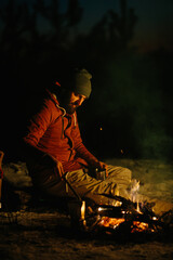 Fototapeta na wymiar A bearded young man rests by the night campfire in the winter forest. The concept of survival and hiking in the wild.