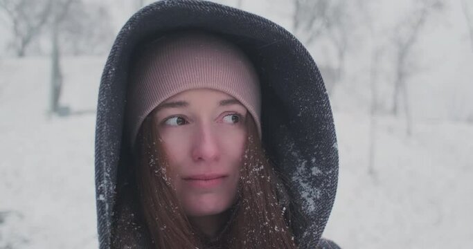 Young caucasian woman in a hood looking at a snow blizzard. Snow gets in her eyes, the girl blinks slowly. Portrait, winter season, cold, frost.
