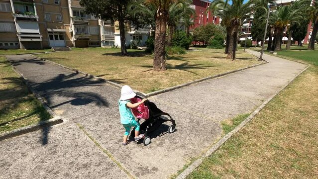 Little girl in a panama rolls a stroller with a toy along the path in the park