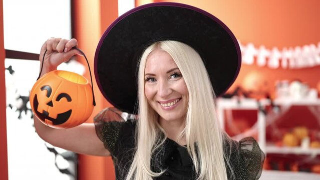 Young blonde woman wearing witch costume holding halloween pumpkin basket at home