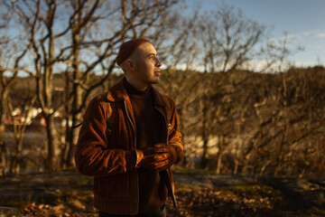 A caucasian man walking in the forest at the top of a hill at a lookout point wearing a leather backpack and a beanie.