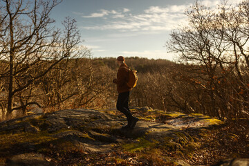 A caucasian man walking in the forest at the top of a hill at a lookout point wearing a leather backpack and a beanie.