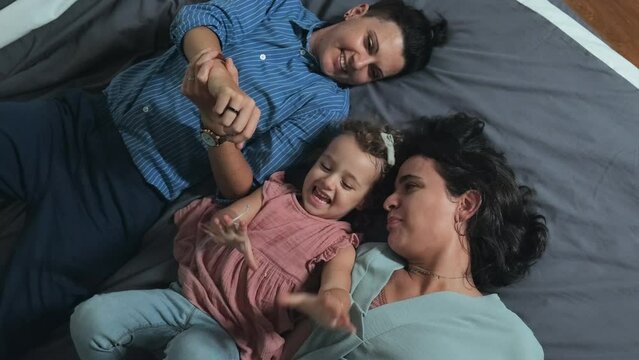 Top view of happy Caucasian lesbian couple and their beloved little daughter playing while lying on bed at home together