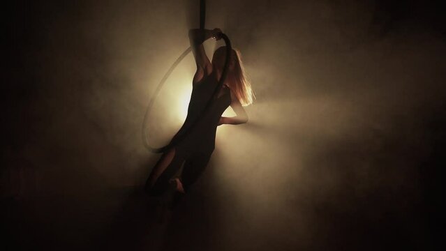 Young woman performs the acrobatic elements in the air hoop. Aerialist in on black background dark studio with backlight. For sports, acrobatic, circus school.