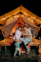 Obraz na płótnie Canvas Happy couple relaxing in glamping on summer evening and drinking wine near cozy bonfire. Luxury camping tent for outdoor recreation and recreation. Lifestyle concept