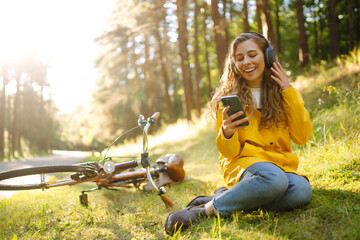 Pretty young womanusing mobile phone in the city while listening music through earphones to go by the bicycle in the park. Lifestyle. Relax, nature concept.