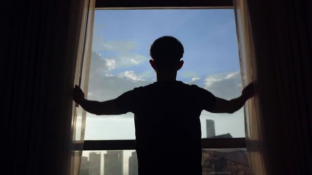 A young man opens the curtains in the morning, raises his arms and stretches. A cinematic man opens large window shades and let the sunlight into the room