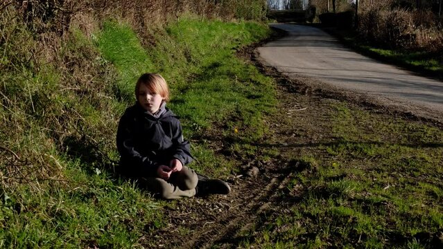 A young boy is sitting on the ground. Tired child from a long walk. A boy of nine years old with long red hair in a warm jacket.