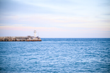 A wide shot captures a white lighthouse at the end of a stone jetty, extending into the calm, blue sea. The horizon is visible, with a soft, pale, overcast sky above.