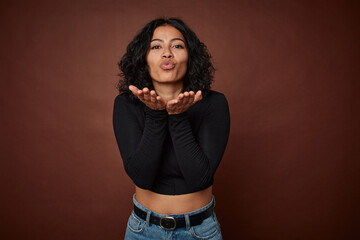 Young colombian curly hair woman isolated on brown background folding lips and holding palms to send air kiss.