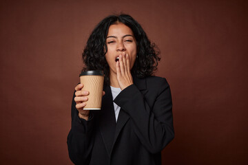 Young businesswoman yawns, holding coffee to-go, tired.