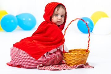 A little girl dressed as a red riding hood sits near a basket. Carnival, Purim, Halloween